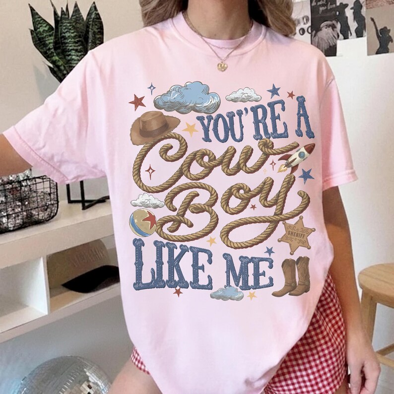 Puede incluir: Una camiseta de manga corta de color rosa claro con un estampado gr&aacute;fico que incluye el texto "YOU'RE A COWBOY LIKE ME" en letras azules. El dise&ntilde;o presenta un sombrero de vaquero marr&oacute;n, una cuerda enrollada, un cohete, una placa de sheriff y botas.