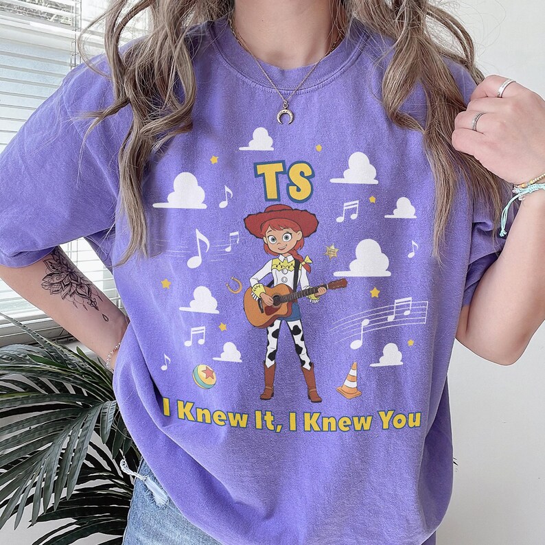 May include: A purple t-shirt featuring a graphic of the character Jessie from Toy Story playing an acoustic guitar. The design includes white clouds, musical notes, a ball, a traffic cone, and the text TS and I Knew It, I Knew You in yellow.