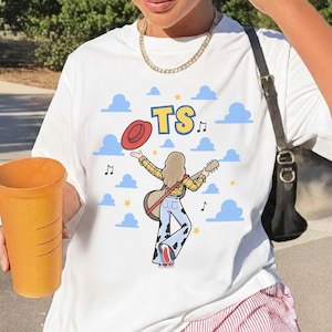 May include: A white t-shirt featuring a graphic of a person with long hair wearing a yellow plaid shirt and cow-print bell-bottom pants, holding a guitar and a red hat. The design includes blue clouds, yellow stars, music notes, and the text TS.