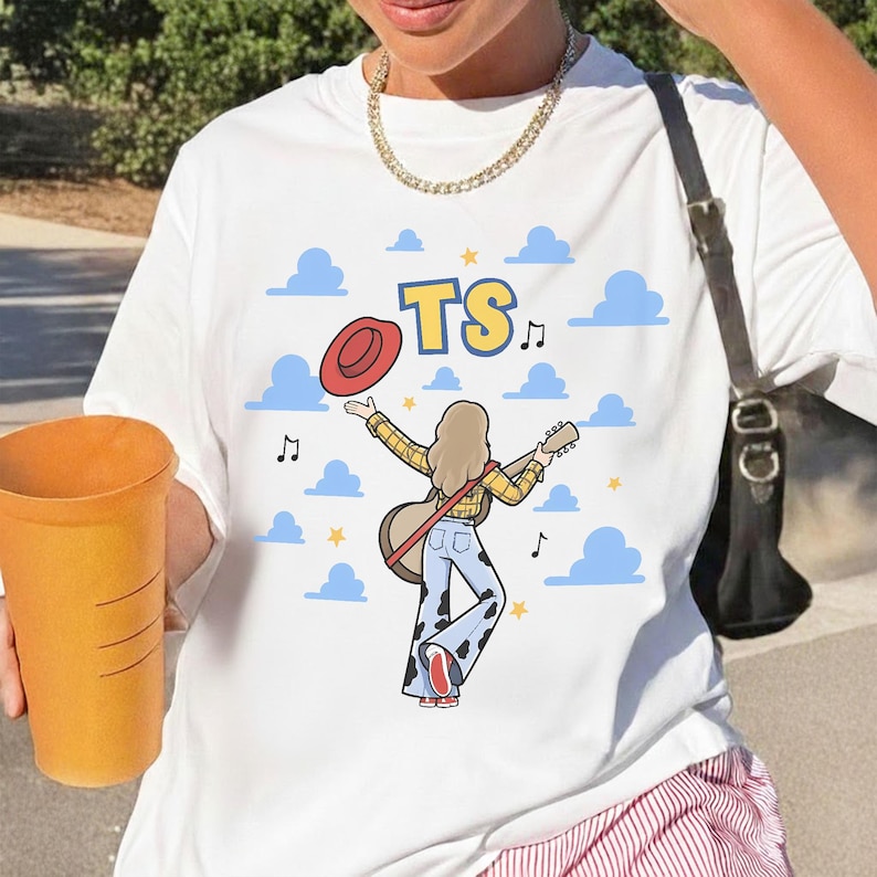 May include: A white t-shirt featuring a graphic of a person with long hair wearing a yellow plaid shirt and cow-print bell-bottom pants, holding a guitar and a red hat. The design includes blue clouds, yellow stars, music notes, and the text TS.