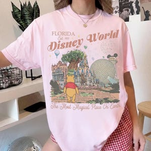 Puede incluir: Camiseta rosa claro con un dise&ntilde;o de Disney World en Florida. El dise&ntilde;o incluye el texto "Florida Est. 1971 Disney World" y una ilustraci&oacute;n de Winnie the Pooh. La frase "The Most Magical Place On Earth" tambi&eacute;n est&aacute; impresa.
