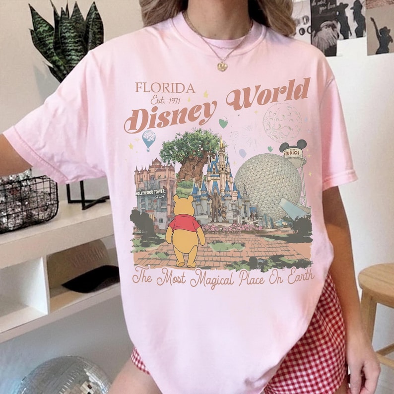 Puede incluir: Camiseta rosa claro con un dise&ntilde;o de Disney World en Florida. El dise&ntilde;o incluye el texto "Florida Est. 1971 Disney World" y una ilustraci&oacute;n de Winnie the Pooh. La frase "The Most Magical Place On Earth" tambi&eacute;n est&aacute; impresa.