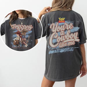 Puede incluir: Una camiseta gris carb&oacute;n con un gr&aacute;fico de Toy Story. La parte posterior muestra el texto 'You're a Cowboy like me' en letra grande sobre un trazo azul. El recuadro frontal muestra dos personajes con guitarras y el texto 'Toy Story'.