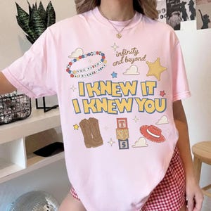 May include: A light pink short-sleeve t-shirt featuring a graphic print with the text "I KNEW IT I KNEW YOU", "infinity and beyond", and "YOU'VE GOT A FRIEND IN ME". The design includes illustrations of a beaded bracelet, cowboy boots, blocks, and a hat.