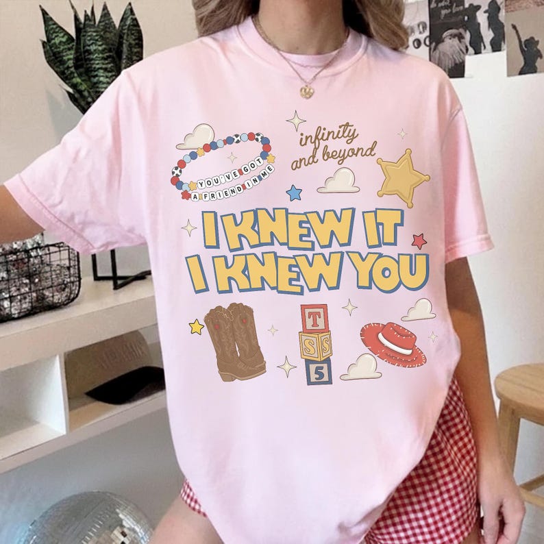 May include: A light pink short-sleeve t-shirt featuring a graphic print with the text "I KNEW IT I KNEW YOU", "infinity and beyond", and "YOU'VE GOT A FRIEND IN ME". The design includes illustrations of a beaded bracelet, cowboy boots, blocks, and a hat.