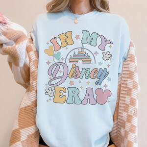 Op de afbeelding: Lichtblauw sweatshirt met de tekst "IN MY Disney ERA" in kleurrijke retroletters. Het ontwerp bevat een kasteel, Mickey Mouse-handschoenen en ballonnen. Een casual top voor Disney-fans.