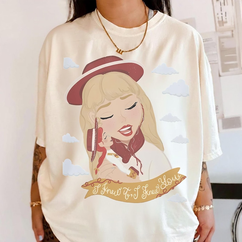 May include: An off-white t-shirt featuring a graphic illustration of a blonde person wearing a maroon hat, holding a small doll with a matching hat. The design includes light blue clouds and a tan banner with the text I Knew It, I Knew You.