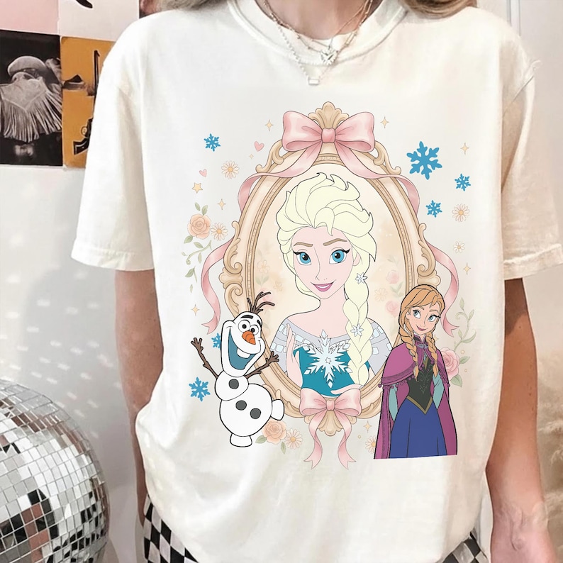 Pu&ograve; includere: T-shirt color crema con un ritratto incorniciato di Elsa di Frozen, con un fiocco rosa e dettagli floreali. Sono raffigurati anche Olaf e Anna. Il design include fiocchi di neve e cuori.