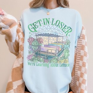 Puede incluir: Camiseta azul claro con un dise&ntilde;o gr&aacute;fico caprichoso. Muestra una estructura tipo carrusel con el texto "GET IN LOSER" y "We're Learning About Lettuce". El dise&ntilde;o incorpora tonos verdes, morados y rosas, creando una est&eacute;tica fantasiosa.