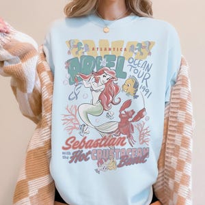 May include: Light blue t-shirt featuring a graphic of Ariel from The Little Mermaid, with the text "Atlantica Ocean Tour 1991." The design includes Ariel, Flounder, Sebastian, and other sea creatures. The shirt has a crew neck.