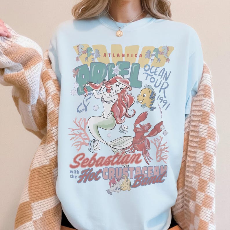 May include: Light blue t-shirt featuring a graphic of Ariel from The Little Mermaid, with the text "Atlantica Ocean Tour 1991." The design includes Ariel, Flounder, Sebastian, and other sea creatures. The shirt has a crew neck.