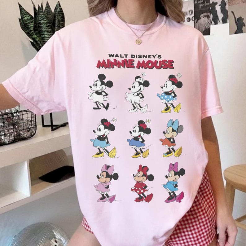 K&ouml;nnte beinhalten: Hellrosa T-Shirt mit einem Aufdruck von Minnie Mouse in verschiedenen Outfits. Der Text "Walt Disney's Minnie Mouse" ist in Rot und Schwarz &uuml;ber den Bildern gedruckt. Das T-Shirt hat kurze &Auml;rmel und einen Rundhalsausschnitt.