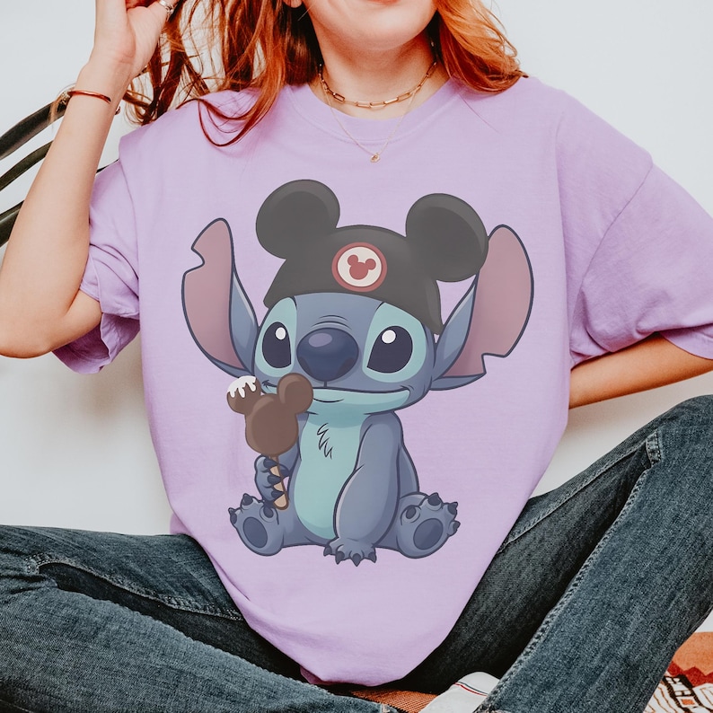 May include: A light purple t-shirt featuring a cartoon illustration of Stitch from Lilo & Stitch. Stitch is wearing a black Mickey Mouse hat and holding a Mickey Mouse ice cream bar. The shirt is a relaxed fit.