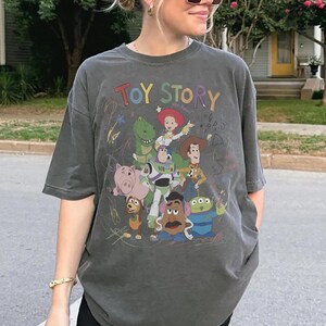 May include: Dark gray t-shirt featuring the words "TOY STORY" in colorful letters above a graphic of characters from the animated film. The design includes Woody, Buzz Lightyear, Rex, and other beloved characters.