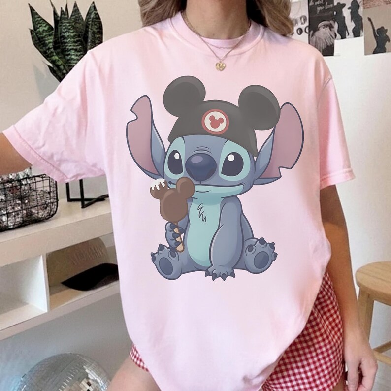 May include: A light pink t-shirt featuring a cartoon illustration of Stitch from Lilo & Stitch. Stitch is wearing a black Mickey Mouse hat and holding a brown ice cream bar. The shirt is a crew neck style.