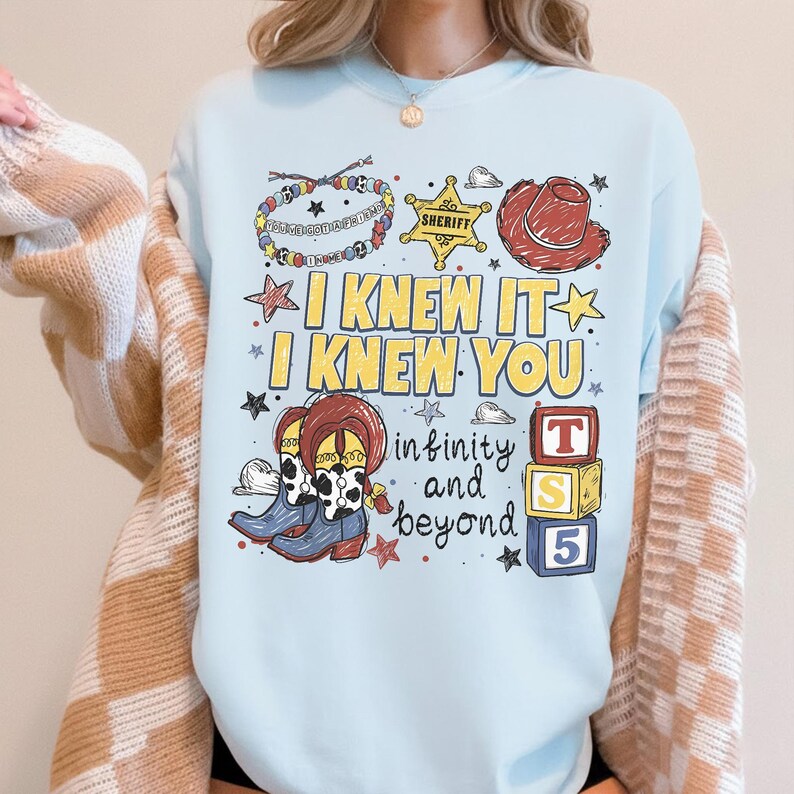 May include: A light blue crewneck sweatshirt featuring a graphic print with the text I KNEW IT I KNEW YOU, infinity and beyond, and T S 5. The design includes illustrations of a beaded bracelet, a sheriff badge, a cowboy hat, boots, and blocks.