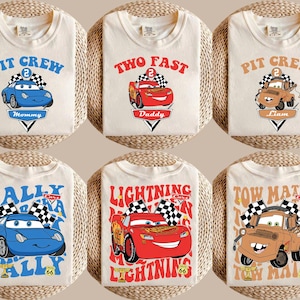 May include: Off-white t-shirts featuring cartoon car designs. The shirts display phrases like "Pit Crew" and "Two Fast", alongside car illustrations in blue, red, and brown. Each shirt has a name or title.