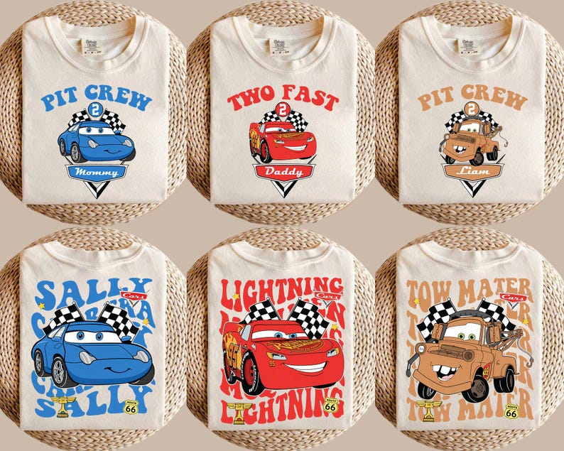 May include: Off-white t-shirts featuring cartoon car designs. The shirts display phrases like "Pit Crew" and "Two Fast", alongside car illustrations in blue, red, and brown. Each shirt has a name or title.