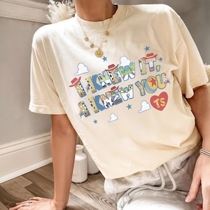 May include: A cream-colored short-sleeve t-shirt featuring the text I KNEW IT, I KNEW YOU in blue and yellow block letters. The letters contain patterns of cowboy and space ranger characters. Includes small blue stars, white clouds, and a red heart.