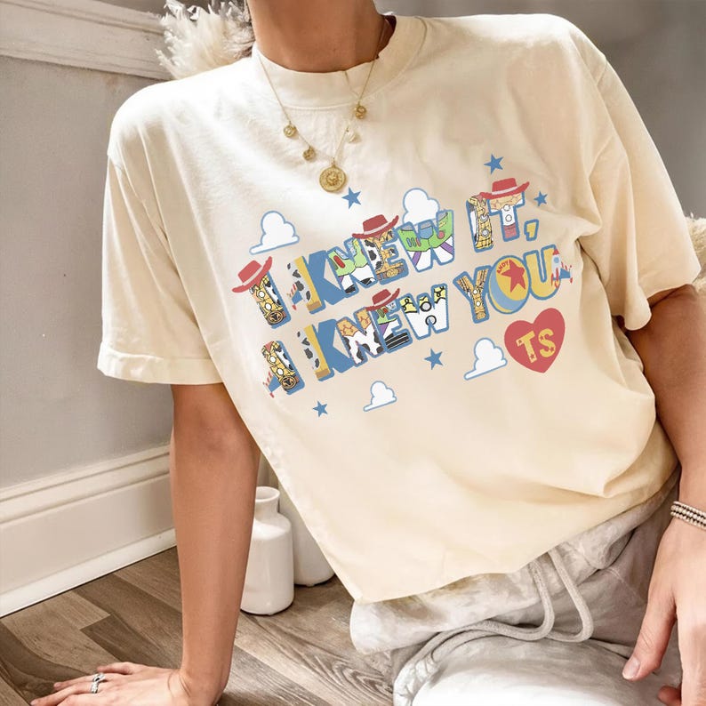 May include: A cream-colored short-sleeve t-shirt featuring the text I KNEW IT, I KNEW YOU in blue and yellow block letters. The letters contain patterns of cowboy and space ranger characters. Includes small blue stars, white clouds, and a red heart.