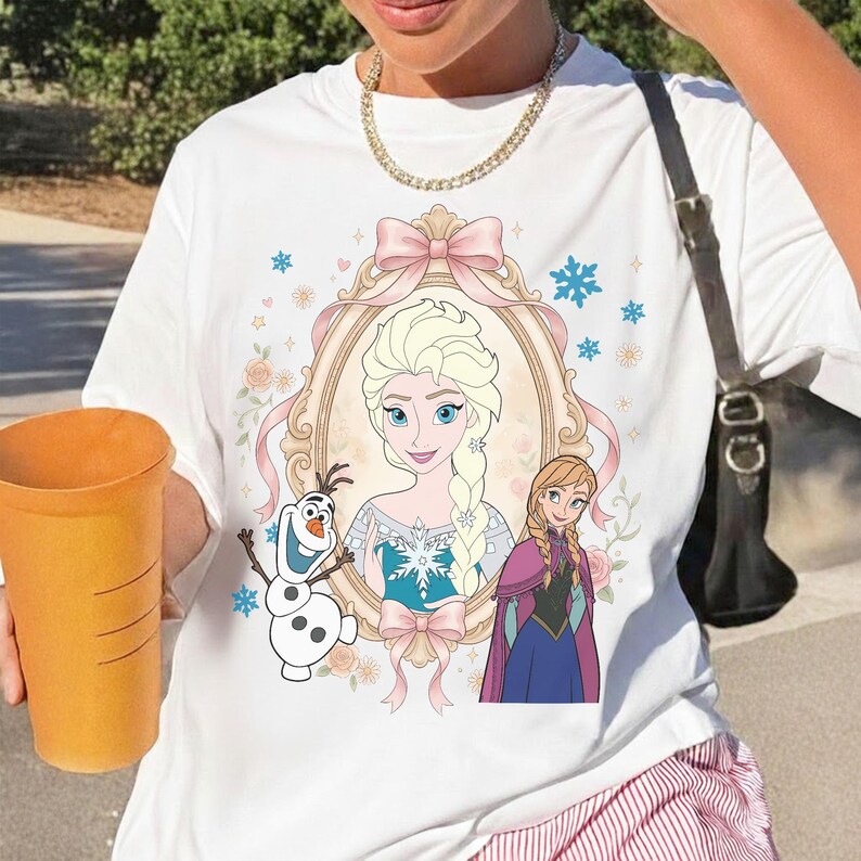 Pu&ograve; includere: T-shirt bianca con un'illustrazione a cartoni animati di Elsa e Anna del film Frozen. Il design include un ritratto di Elsa in una cornice decorativa, con Olaf il pupazzo di neve e Anna. Fiocco rosa e accenti floreali completano il design.