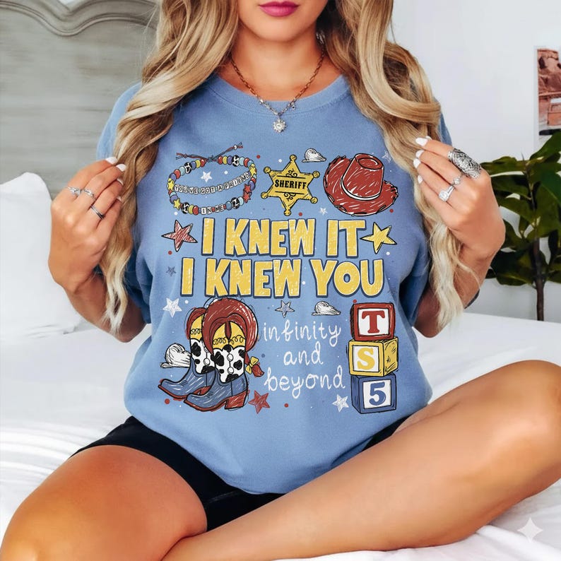 May include: A light blue t-shirt featuring a graphic print with the text I KNEW IT I KNEW YOU infinity and beyond. The design includes a sheriff star, a cowboy hat, cow print boots, a beaded bracelet, and three stacked blocks labeled T S 5.