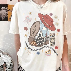 May include: A cream-colored short-sleeve t-shirt featuring a graphic print of a cow-print cowboy boot, a red cowboy hat, stacked blocks with the letters T, S, and a number 13, a bow, flowers, and a beaded string with the text I KNEW IT.