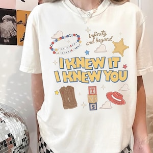 May include: A cream-colored short-sleeve t-shirt featuring a graphic print with the text "I KNEW IT I KNEW YOU", "infinity and beyond", and "YOU'VE GOT A FRIEND IN ME". The design includes illustrations of a beaded bracelet, a sheriff star, cowboy boots, alphabet blocks, and a red hat.