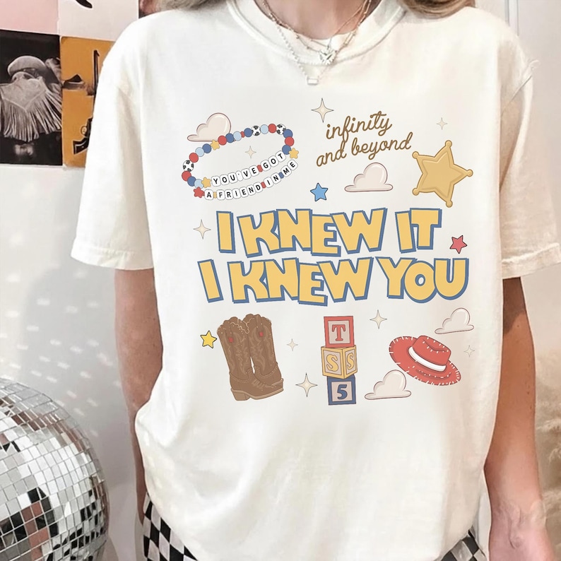May include: A cream-colored short-sleeve t-shirt featuring a graphic print with the text "I KNEW IT I KNEW YOU", "infinity and beyond", and "YOU'VE GOT A FRIEND IN ME". The design includes illustrations of a beaded bracelet, a sheriff star, cowboy boots, alphabet blocks, and a red hat.