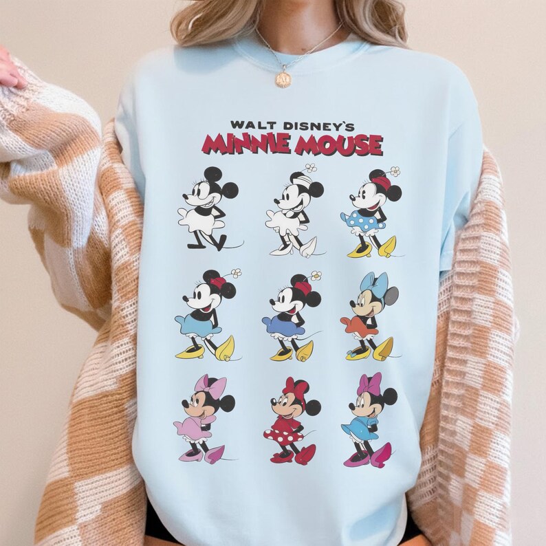 K&ouml;nnte beinhalten: Hellblaues Sweatshirt mit einem Aufdruck von Minnie Mouse in verschiedenen Outfits. Der Text "WALT DISNEY'S MINNIE MOUSE" ist in Rot und Schwarz &uuml;ber den Illustrationen gedruckt. Das Design enth&auml;lt mehrere Bilder von Minnie Mouse.