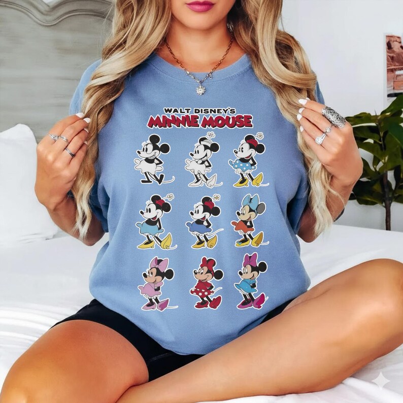 K&ouml;nnte beinhalten: Hellblaues T-Shirt mit einem Aufdruck von Minnie Mouse in verschiedenen Outfits. Der Text "WALT DISNEY'S MINNIE MOUSE" ist oben aufgedruckt. Das Design zeigt Minnie Mouse in verschiedenfarbigen Kleidern und Schleifen.