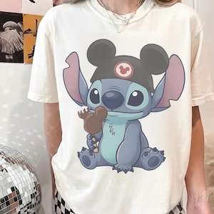 May include: Cream-colored t-shirt featuring a cartoon illustration of Stitch wearing a Mickey Mouse hat and holding a Mickey Mouse-shaped ice cream bar. The character is blue with large ears and eyes. The shirt is casual wear.