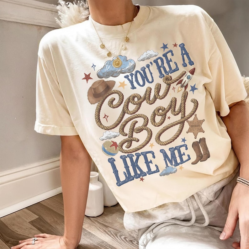 Puede incluir: Una camiseta de manga corta color crema con un estampado gr&aacute;fico que incluye el texto "YOU'RE A COWBOY LIKE ME" en azul. El dise&ntilde;o presenta un sombrero de vaquero marr&oacute;n, una cuerda, un cohete, una estrella de sheriff, una pelota y nubes.