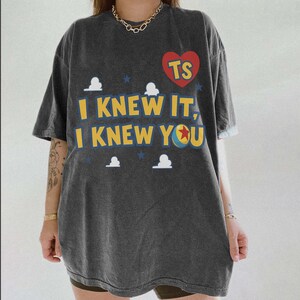 May include: A charcoal gray short-sleeve t-shirt featuring a red heart with the letters TS, yellow text reading I KNEW IT, I KNEW YOU, small white cloud shapes, blue stars, and a colorful ball graphic with a red star in the center of the text.
