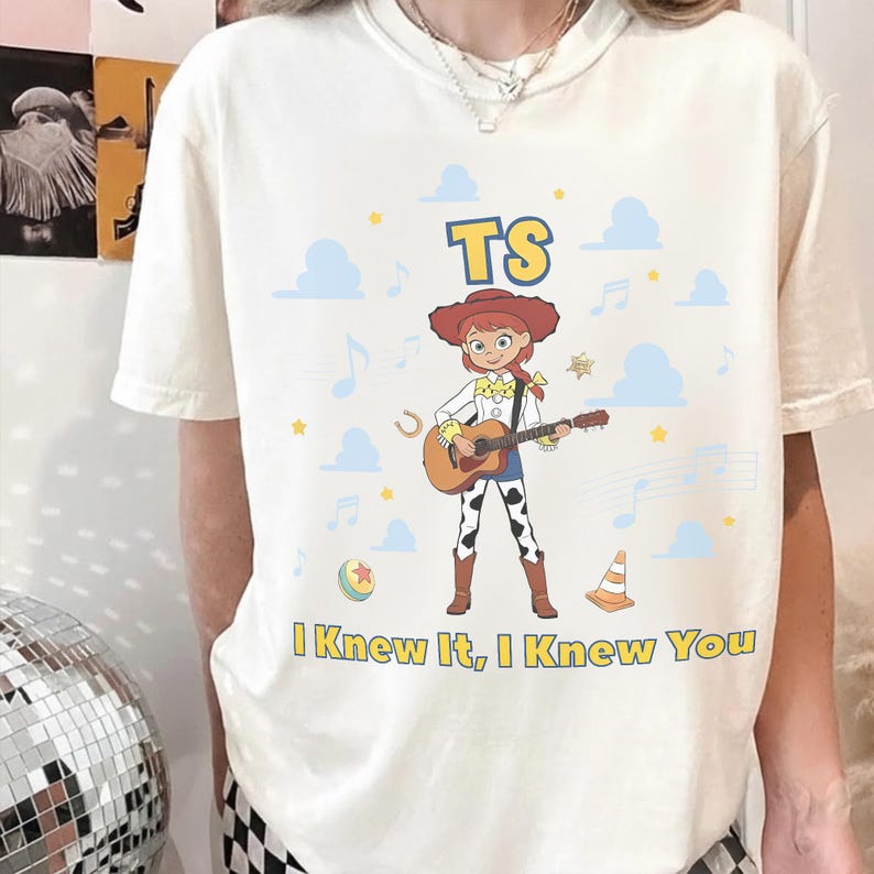 May include: A cream-colored t-shirt featuring a graphic of a cowgirl character playing an acoustic guitar. The design includes the text TS, musical notes, clouds, a ball, and a traffic cone, with the text I Knew It, I Knew You at the bottom.
