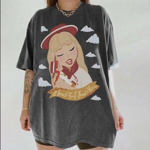 May include: A charcoal grey short-sleeve t-shirt featuring a graphic illustration of a blonde character wearing a red hat, holding a small doll, surrounded by white clouds. A gold banner at the bottom displays the text, I Knew It. I Knew You.