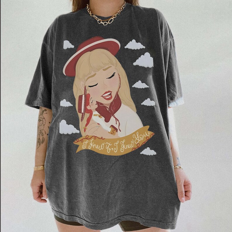 May include: A charcoal grey short-sleeve t-shirt featuring a graphic illustration of a blonde character wearing a red hat, holding a small doll, surrounded by white clouds. A gold banner at the bottom displays the text, I Knew It. I Knew You.