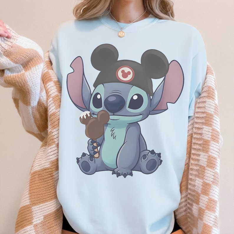 May include: Light blue sweatshirt featuring a cartoon illustration of Stitch wearing a Mickey Mouse hat and holding a chocolate ice cream bar. The graphic is centered on the front of the shirt. The sweatshirt is a solid light blue color.