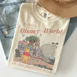 Puede incluir: Camiseta color crema con un dise&ntilde;o de Disney World en Florida. El dise&ntilde;o incluye el texto "FLORIDA Est. 1971 Disney World" y presenta ilustraciones de lugares emblem&aacute;ticos del parque y Winnie the Pooh. La camiseta est&aacute; doblada sobre unos vaqueros azules.