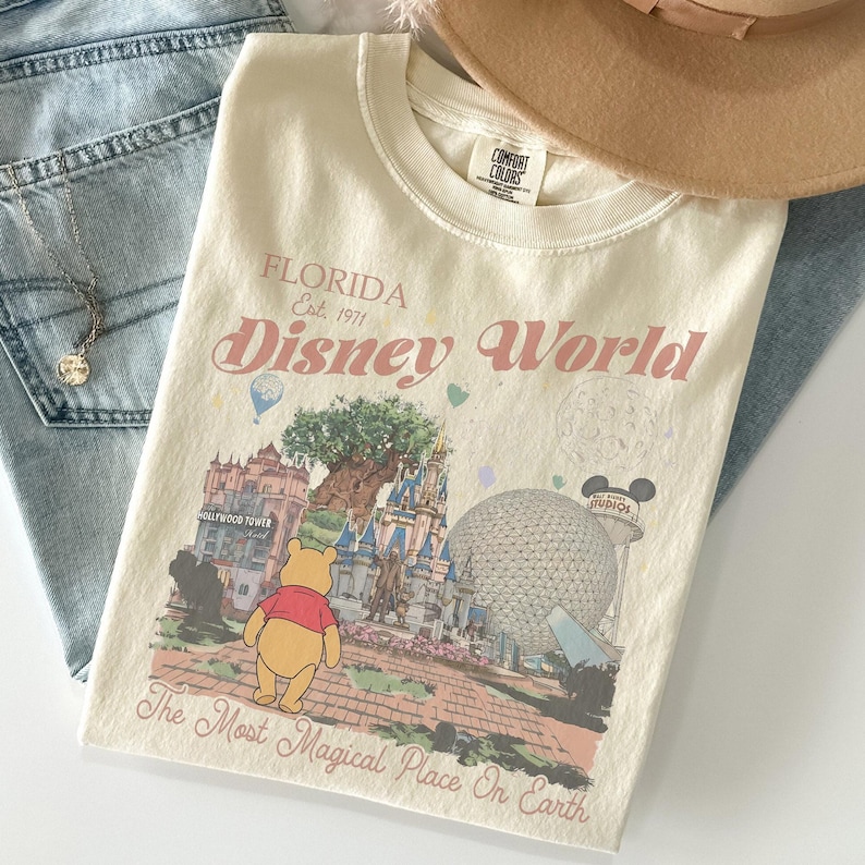 Puede incluir: Camiseta color crema con un dise&ntilde;o de Disney World en Florida. El dise&ntilde;o incluye el texto "FLORIDA Est. 1971 Disney World" y presenta ilustraciones de lugares emblem&aacute;ticos del parque y Winnie the Pooh. La camiseta est&aacute; doblada sobre unos vaqueros azules.