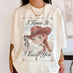 May include: A cream-colored t-shirt featuring a graphic of a cartoon cowgirl wearing a brown hat and a cow-print glove. The text reads "EST 2026", "I Knew It", and "I Knew You" with a musical note symbol. The design is framed by a rope-like border.