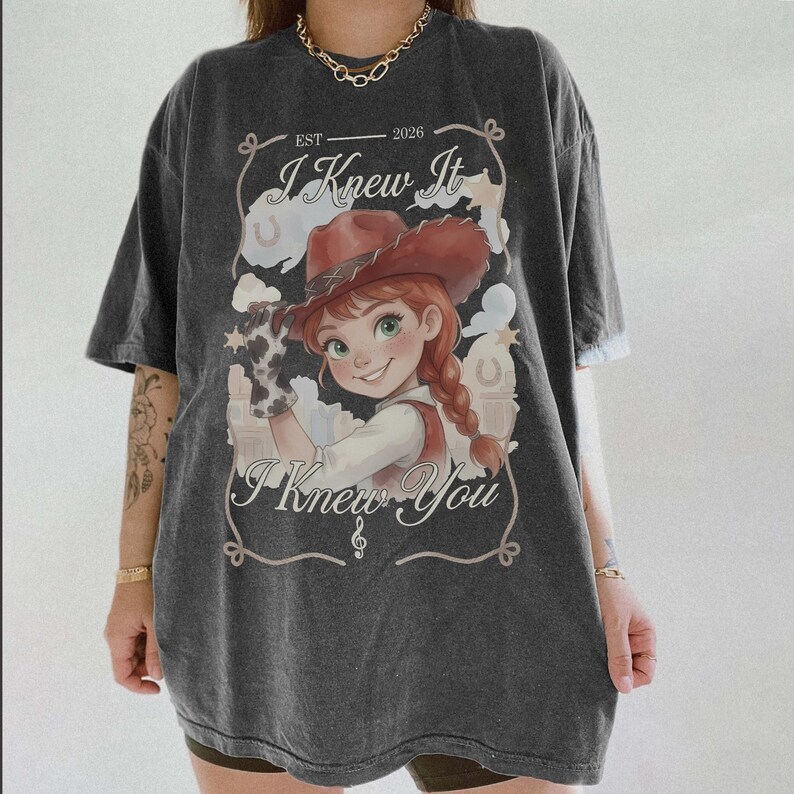 May include: A charcoal gray oversized t-shirt featuring a graphic of a cartoon character wearing a brown cowboy hat and a cow-print glove. The text reads "EST 2026", "I Knew It", and "I Knew You" with decorative line art and stars.