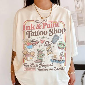 May include: Cream-colored t-shirt featuring a colorful graphic for a tattoo shop. The design includes various illustrations like a skull, a boot with a snake, and a boat. Text reads "Magic's Ink & Paint Tattoo Shop" and "The Most Magical Tattoos on Earth!"