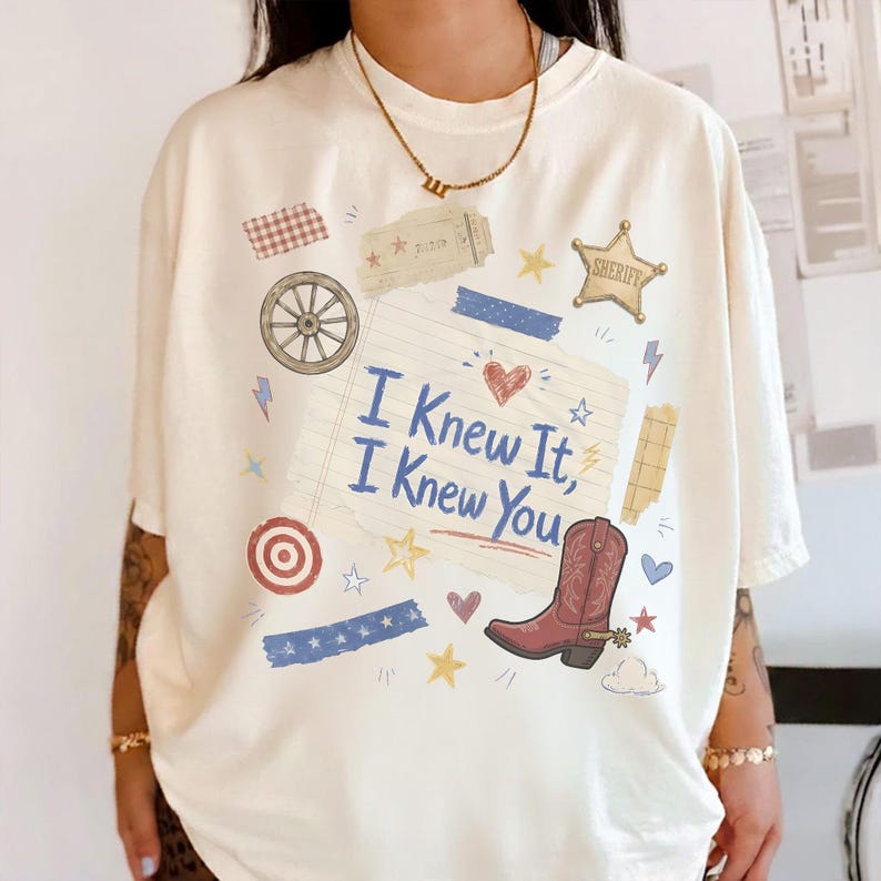 May include: An off-white short-sleeve t-shirt featuring a graphic print of a lined paper note with the text I Knew It, I Knew You, surrounded by illustrations of a wagon wheel, a sheriff star, a red cowboy boot, a target, and various stars.