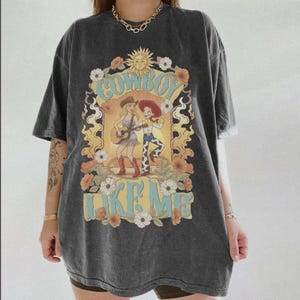 May include: A charcoal grey short-sleeve t-shirt featuring a graphic print of two cowgirls, one playing a guitar, surrounded by flowers and a sun. The text "COWBOY LIKE ME" is printed in light blue letters above and below the central illustration.
