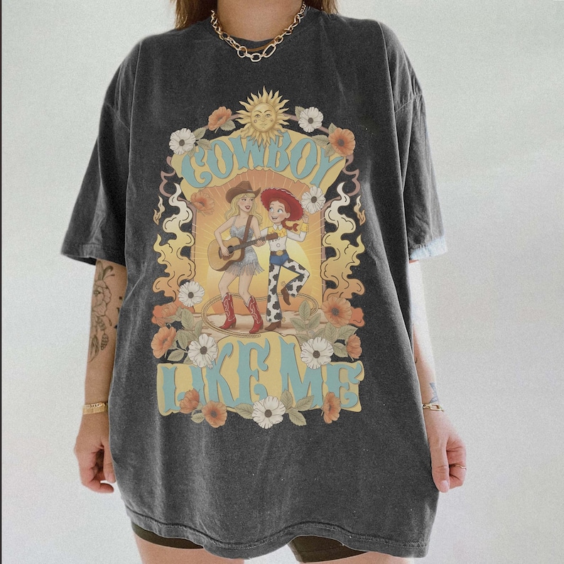 May include: A charcoal grey short-sleeve t-shirt featuring a graphic print of two cowgirls, one playing a guitar, surrounded by flowers and a sun. The text "COWBOY LIKE ME" is printed in light blue letters above and below the central illustration.