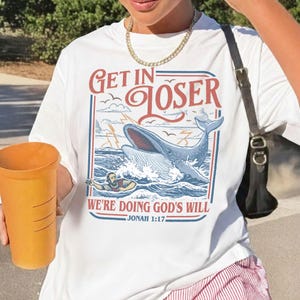K&ouml;nnte beinhalten: Wei&szlig;es T-Shirt mit einem grafischen Design, das einen Wal zeigt, der aus dem Wasser springt, eine Person im Wasser und den Text "GET IN LOSER WE'RE DOING GOD'S WILL" mit "Jonah 1:17" darunter. Das Design ist rot, blau und wei&szlig;.