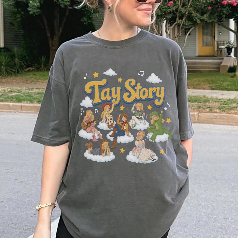 May include: A charcoal gray t-shirt featuring the text Tay Story in yellow letters. The graphic includes multiple illustrated figures dressed in various outfits, each positioned on white clouds surrounded by yellow stars and musical notes.