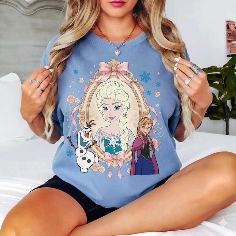 Pu&ograve; includere: T-shirt azzurra con un ritratto di Elsa di Frozen, incorniciato da un fiocco rosa e dettagli floreali. Il design include immagini di Olaf e Anna. La maglietta &egrave; un capo comodo e casual.