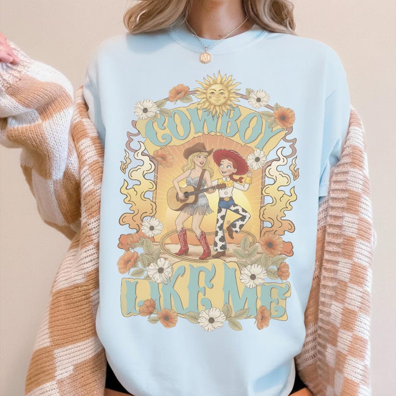 May include: A light blue crewneck sweatshirt featuring a graphic print of two figures in cowboy hats, one playing an acoustic guitar. The design includes a sun, flowers, and the text "COWBOY LIKE ME" in a stylised font, framed by swirling decorative lines.